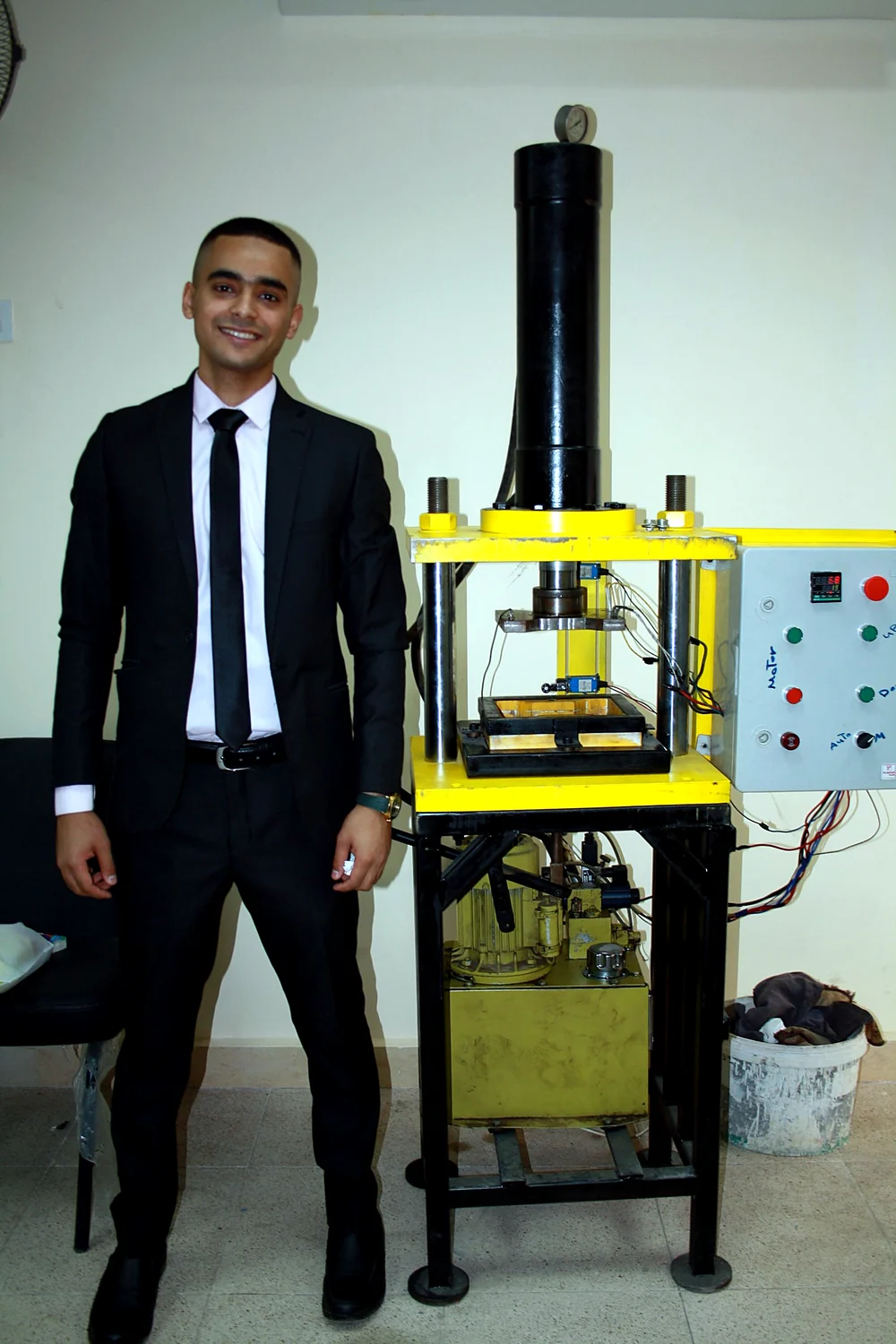 Ahmed standing next to his rubber recycling machine, a yellow hydraulic press with a control panel